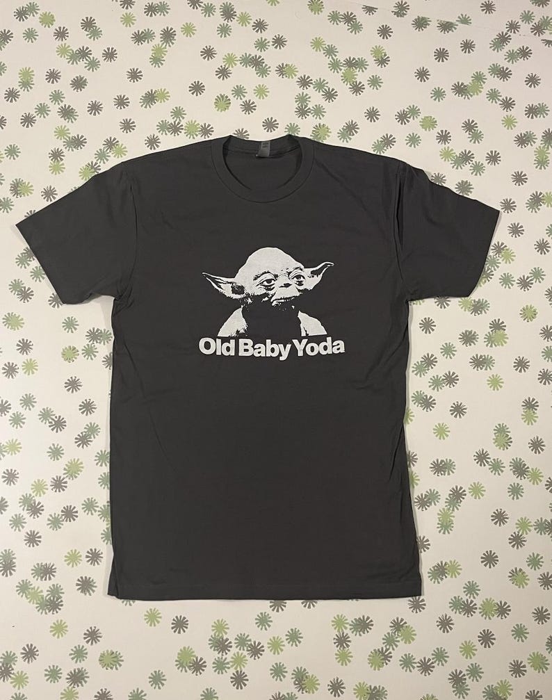May include: A dark gray t-shirt with a white graphic of Yoda from Star Wars. The text "Old Baby Yoda" is printed below the image.