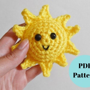 May include: A crocheted yellow sun with a smiling face and black eyes. The sun is being held in a hand. The text "PDF Pattern" is in a blue circle.