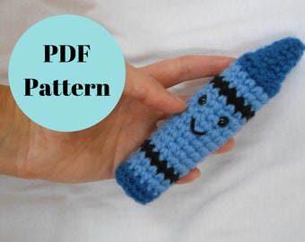 May include: A blue crocheted crayon with a black stripe and a smiling face. The crayon is being held in a hand. PDF Pattern.