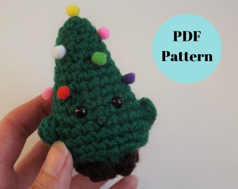 Könnte beinhalten: Ein grüner gehäkelter Weihnachtsbaum mit einem lächelnden Gesicht und bunten Ornamenten. Der Baum wird in einer Hand gehalten. PDF-Muster.