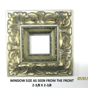 May include: A square picture frame with an ornate, embossed scrollwork pattern in a metallic gold and dark grey finish. The frame features a central opening with dimensions clearly printed as 2-3/8 inches by 2-3/8 inches on the front.