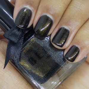 May include: A bottle of black nail polish with gold glitter. The bottle is labelled "BE" and "HANDMADE IN THE UK".