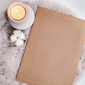Op de afbeelding: Een printable PDF slaaptracker-blad op bruin papier met een raster van nummers van 1 tot 30. Het blad ligt op een lichtbruin, pluche tapijt met een witte kaarshouder en een wattenbol.