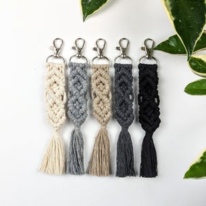 May include: Five macrame keychains with tassels in various colours, including beige, grey, and black. The keychains are hanging from silver key rings.
