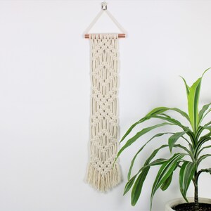 May include: A white macrame wall hanging with a geometric pattern and a wooden dowel rod at the top. The hanging is attached to a white wall with a small metal hook. A green plant is in a pot in the lower right corner of the image.