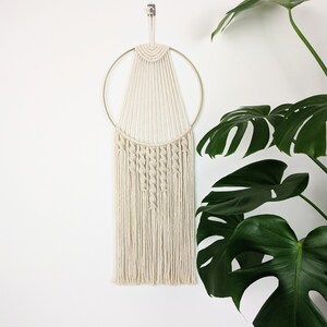Peut inclure: Une suspension murale en macramé blanche avec un cercle doré et des franges. La suspension est fixée à un mur blanc avec une plante en arrière-plan.