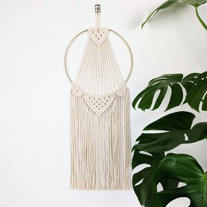 Peut inclure: Une suspension murale en macramé blanche avec un cercle doré et des franges. La suspension a la forme d'un triangle avec un motif géométrique.