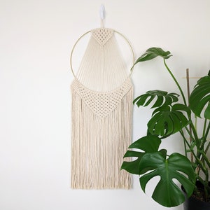 May include: A macrame wall hanging with a gold hoop and a geometric design. The hanging is made with natural coloured yarn and has a fringe at the bottom.