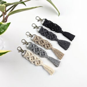 May include: Five macrame keychains with silver key rings and tassels. The keychains are in shades of grey, beige, and black.