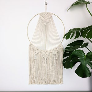 Peut inclure: Une suspension murale en macramé blanche avec un cercle en métal doré et des franges. La suspension est réalisée avec une technique de nouage et présente un motif géométrique.