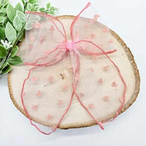 May include: A pink bow made of sheer fabric adorned with small heart-shaped accents. The bow is centered on a wooden slice, with green foliage in the background. The bow's edges are outlined with a darker pink trim.