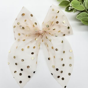 May include: A large, blush-colored tulle bow with gold polka dots. The bow is tied in a knot in the center and has two long tails.