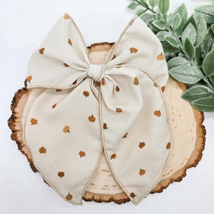 May include: A large cream-colored bow with a repeating pattern of small orange pumpkins. The bow is centered on a wooden slice with a natural bark edge, suggesting a fall or harvest theme. The bow is tied in the center.