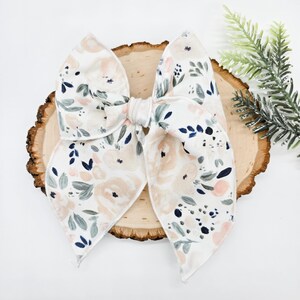 Op de afbeelding: Een witte strik van stof met een bloemenpatroon in tinten roze, groen en blauw. De strik is in een knoop geknoopt en ligt op een houten oppervlak.