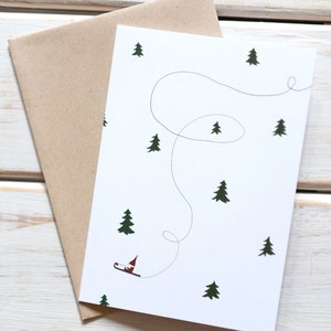 Könnte beinhalten: Eine weiße Grußkarte mit einer einfachen Illustration einer kleinen Figur auf einem Schlitten, die einen gewundenen Weg hinunterfährt. Der Hintergrund ist ein weißes Feld mit kleinen grünen Bäumen, die verstreut sind. Die Karte wird mit einem braunen Umschlag geliefert. Der Text auf der Karte lautet "Tomte on Sled Greeting Card".