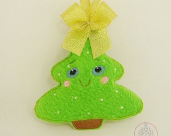 Könnte beinhalten: Ein grüner Weihnachtsbaumschmuck aus Filz mit einem lächelnden Gesicht, blauen Augen und rosa Wangen. Der Baum hat einen braunen Sockel und ist mit kleinen weißen Sternen verziert. Oben ist ein goldenes Band und eine Schleife zum Aufhängen befestigt.