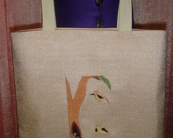 May include: A beige canvas tote bag with a brown tree and birds embroidered on the front. The tree has a hollow trunk with pink birds inside. The birds on the branches are brown and yellow.