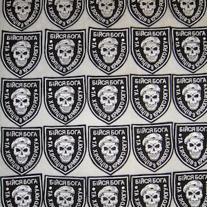 Puede incluir: Veinticinco parches bordados en blanco y negro con un diseño de calavera y huesos cruzados. Los parches tienen forma de escudo y tienen el texto "БІЙСЯ БОГА" y "ТА ХЛОПЦІВ З КРИВОГО РОГУ" en cirílico.