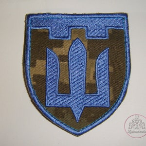以下が含まれることがあります： 迷彩柄の背景に、三叉のシンボルが刺繍された青いパッチ。パッチは盾の形をしています。