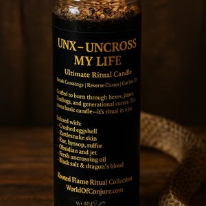 K&ouml;nnte beinhalten: Eine schwarze Ritualkerze in einem Glas mit brennendem Docht. Das Etikett tr&auml;gt den goldenen Schriftzug UNX-UNCROSS MY LIFE, Ultimate Ritual Candle, und listet Inhaltsstoffe wie zersto&szlig;ene Eierschalen, Schlangenhaut, Raute, Ysop, Schwefel, Obsidian, Gagat, Entzauberungs&ouml;l, schwarzes Salz und Drachenblut auf.