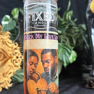 May include: A tall cylindrical glass candle featuring a label with the text "FIXED CANDLE by Shannon Hajira" and "UNBLOCK MY LOVE LIFE". The label depicts a person holding a heart-shaped padlock with a key, with text below reading "Draws a loyal mate fast" and "Opens your love roads".