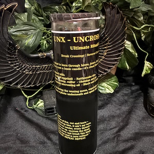 K&ouml;nnte beinhalten: Eine hohe, zylindrische schwarze Kerze in einem Glasbeh&auml;lter mit einem Etikett, das den gelben Text UNX - UNCROSS Ultimate Ritual tr&auml;gt. Das Etikett enth&auml;lt Anweisungen und Zutatenlisten vor einem dunklen Hintergrund mit schwarzen Fl&uuml;geln.