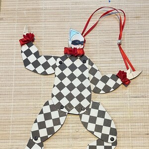 May include: Wooden harlequin ornament with a black and white checkered pattern. The ornament has red ribbon accents on the collar, wrists, and feet. A red ribbon is attached for hanging.