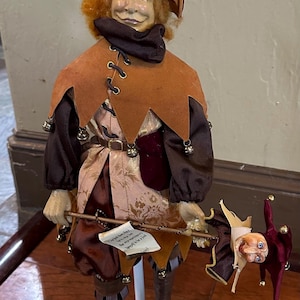 May include: A jester doll with orange hair, wearing a brown velvet hat with bells, a brown suede-like collar, and a patterned tunic. The doll holds a staff topped with a miniature jester head and a small scroll with the text The fool doth think he is wise, but the wise man knows himself to be a fool.