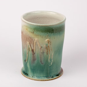 May include: A green, brown, and white ceramic mug with a textured, wavy design. The mug has a slightly uneven rim and a rounded base.