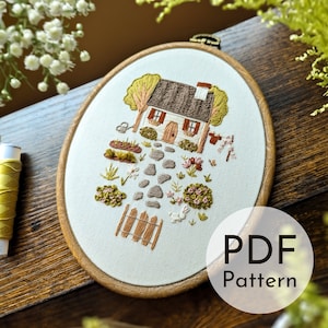 Puede incluir: Arte de bordado en aro que presenta una cabaña blanca con techo marrón, un camino de piedra que conduce a la puerta principal y un pequeño jardín con flores y arbustos. El aro es de madera y tiene un fondo de tela blanca. El texto "PDF Pattern" es visible en la esquina inferior derecha de la imagen.