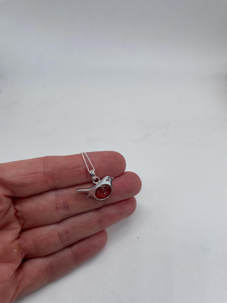 May include: A silver-colored bird pendant necklace with a red, textured center. The bird is small and detailed, with a delicate chain. The pendant is held in a hand against a white background.