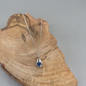 K&ouml;nnte beinhalten: Eine silberne Halskette mit einem tropfenf&ouml;rmigen Anh&auml;nger. Der Anh&auml;nger hat ein blaues Herz im Inneren.