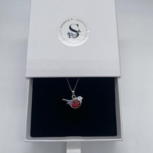 May include: A silver bird pendant with a red center, displayed in an open white gift box with a black interior. The box has a circular sticker with the text "Sparkle & Handmade Gifts". The pendant hangs on a silver chain.