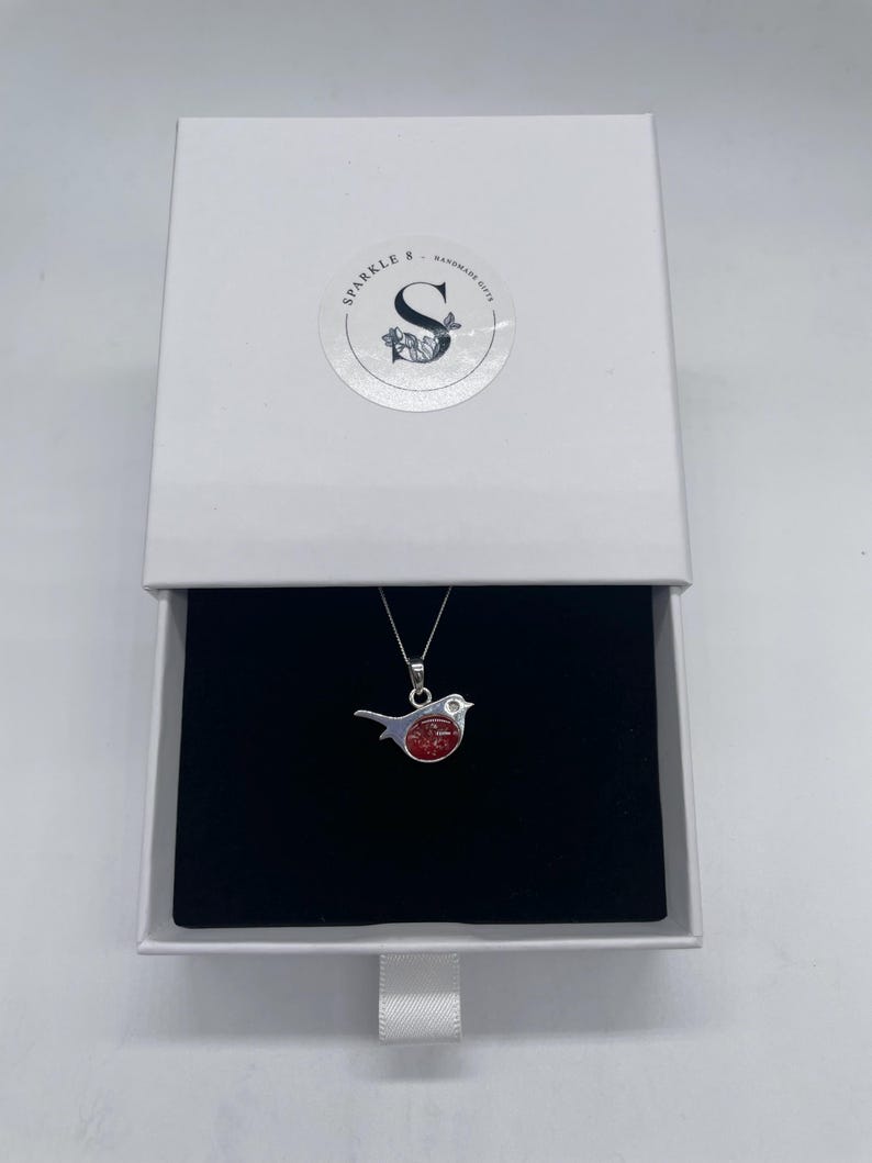 May include: A silver bird pendant with a red center, displayed in an open white gift box with a black interior. The box has a circular sticker with the text "Sparkle & Handmade Gifts". The pendant hangs on a silver chain.