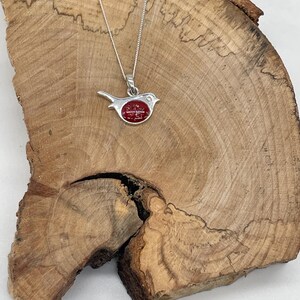 May include: A silver bird pendant necklace with a red center stone. The bird is perched on a silver chain. The necklace is displayed on a wooden surface.