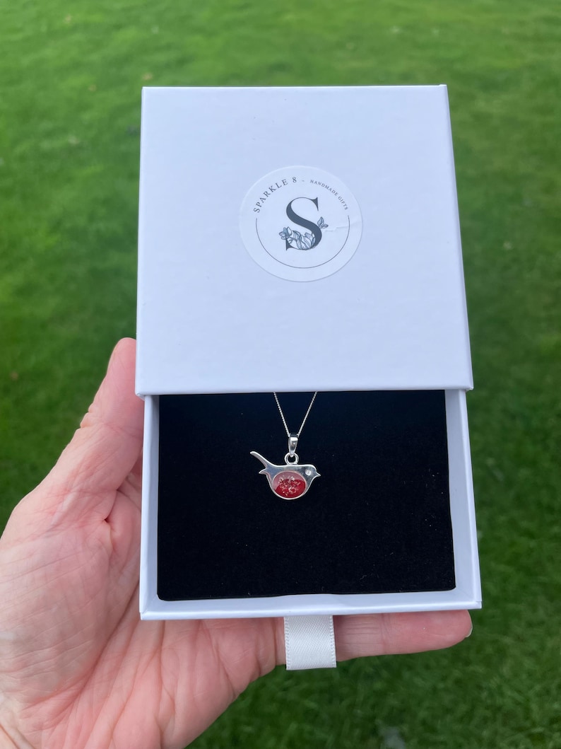 May include: A silver bird pendant necklace with a red and clear center, presented in a white gift box with the text "SPARKLE & HANDMADE GIFTS". The necklace is displayed against a black velvet interior.