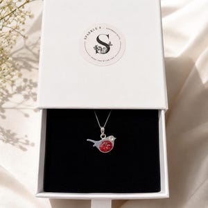 May include: A silver necklace with a red bird pendant inside an open white gift box. The box has a black interior and a ribbon pull. The box lid features a circular sticker with the text "Sparkle & Handmade Gifts" and a floral design.