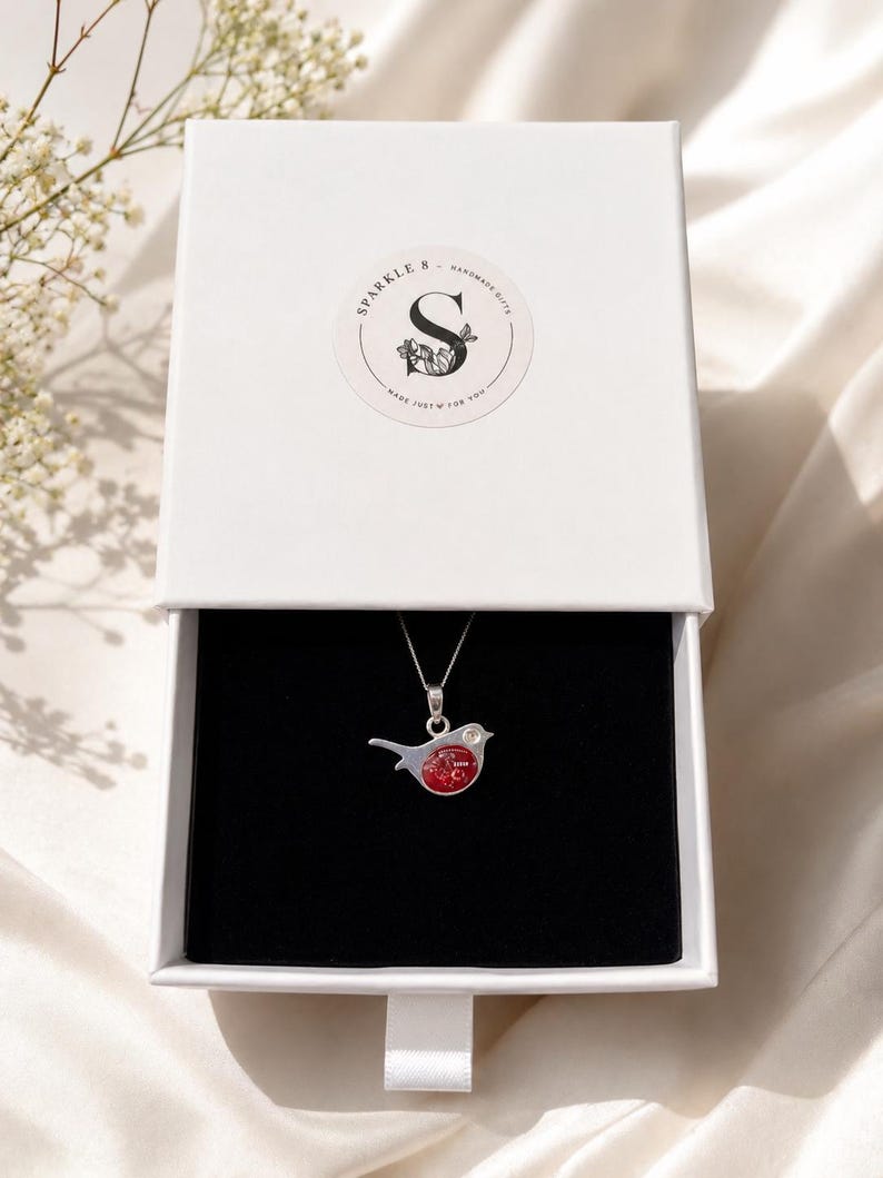 May include: A silver necklace with a red bird pendant inside an open white gift box. The box has a black interior and a ribbon pull. The box lid features a circular sticker with the text "Sparkle & Handmade Gifts" and a floral design.