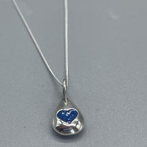 K&ouml;nnte beinhalten: Eine silberne Halskette mit einem tropfenf&ouml;rmigen Anh&auml;nger. Der Anh&auml;nger hat ein blaues Herzdesign im Inneren.