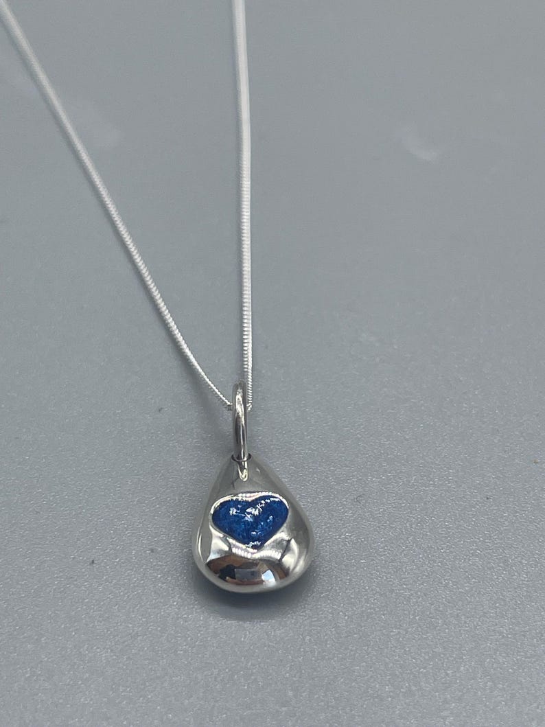 K&ouml;nnte beinhalten: Eine silberne Halskette mit einem tropfenf&ouml;rmigen Anh&auml;nger. Der Anh&auml;nger hat ein blaues Herzdesign im Inneren.