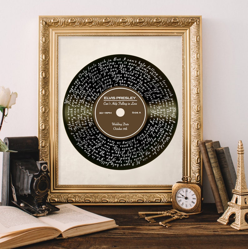 Puede incluir: Una impresi&oacute;n enmarcada de un disco de vinilo negro con la letra de "Can't Help Falling in Love" de Elvis Presley impresa en blanco. La etiqueta del disco es marr&oacute;n con el t&iacute;tulo de la canci&oacute;n, el nombre del artista y la fecha de la boda "19 de octubre".
