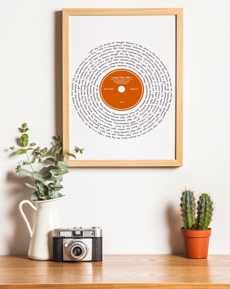 May include: A framed print of a record with the lyrics to the song "Summertime Sadness" by Lana Del Rey. The record is orange and the lyrics are written in black ink.