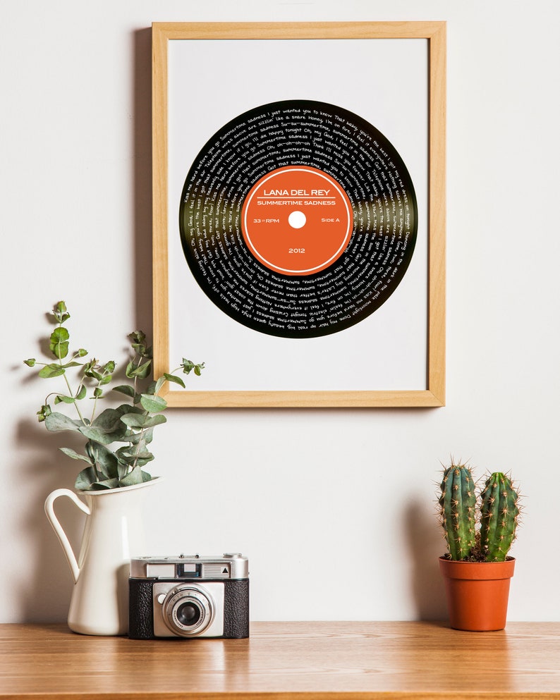 Puede incluir: Una impresi&oacute;n enmarcada de un disco de vinilo negro con el texto "Lana Del Rey Summertime Sadness 33 RPM Side A 2012" en blanco. El disco es naranja y negro con texto blanco.