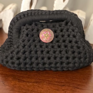 May include: A black crocheted handbag with a rounded shape and a small handle. The bag features a decorative, round button with pink and gold accents. The bag is sitting on a wooden surface.