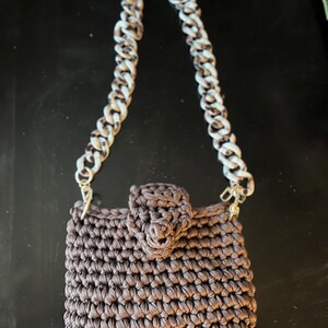 May include: A brown crocheted handbag with a chain strap. The bag has a rectangular shape with a flap closure. The chain strap is a mix of white and brown links, and the bag is made of a tightly woven material.