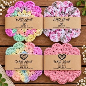 May include: Four round handmade crochet coasters with scalloped edges, each wrapped in a tan paper band labeled White Heart Crochet Set of 4. The coasters feature variegated yarn in pastel pink, yellow, blue, green, and solid pink colorways.