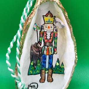 May include: Hand-painted oyster shell Christmas ornament featuring a nutcracker soldier. The ornament has a gold-painted edge and a green and white twisted cord for hanging. The nutcracker is painted in bright colors.