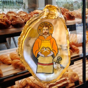 May include: An oyster shell with a gold-colored rim features a hand-painted image of St. Honore, holding bread. The saint is depicted in an orange robe and yellow apron. The background shows various baked goods.