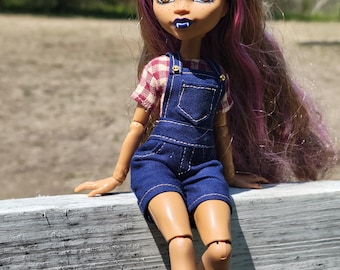 May include: A doll with brown skin, purple and brown hair, and cat ears. The doll wears a red and white plaid shirt under blue denim overalls. The doll has yellow eyes and blue lipstick. The doll is sitting on a white wooden surface.