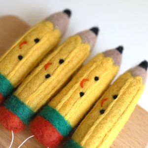 May include: Four felt pencil ornaments with smiling faces. The pencils are yellow with green and red accents.